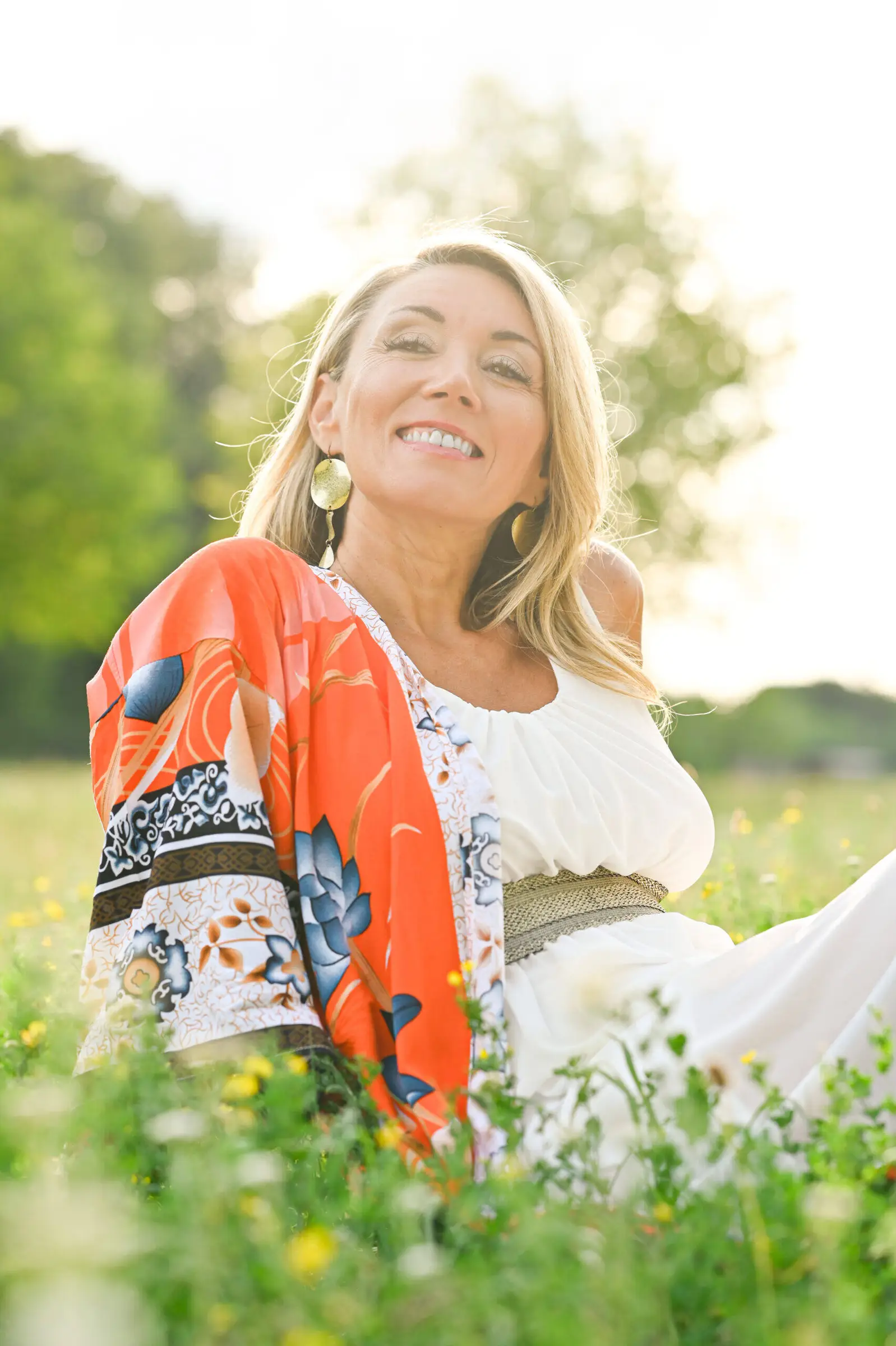 Nelli Halfinger sitzt lachend in einer sonnigen Blumenwiese, weißes Kleid mit orangefarbenem Kimono